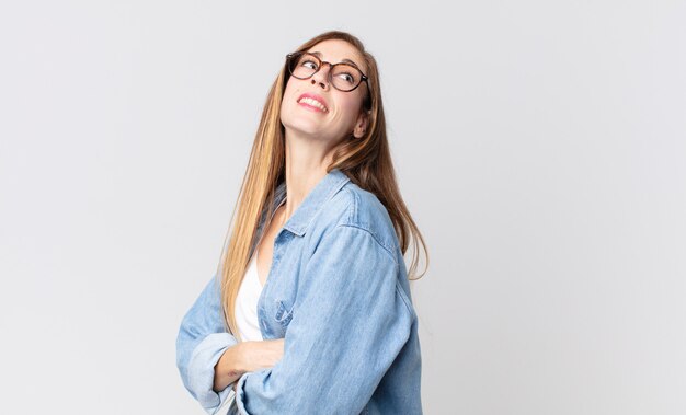 Mulher bonita e magra sorrindo alegremente, sentindo-se feliz, satisfeita e relaxada, de braços cruzados e olhando para o lado