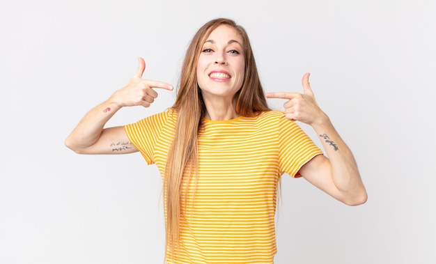 Mulher bonita e magra sorrindo com confiança apontando para o próprio sorriso largo, atitude positiva, relaxada e satisfeita