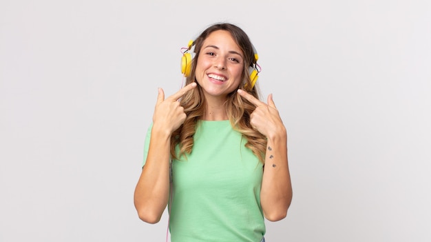 Foto mulher bonita e magra sorrindo com confiança, apontando para o próprio sorriso largo ouvindo música com fones de ouvido