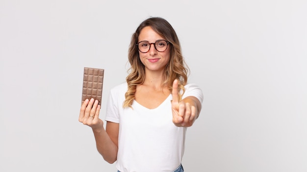 Mulher bonita e magra sorrindo com orgulho e confiança fazendo o número um e segurando uma barra de chocolate