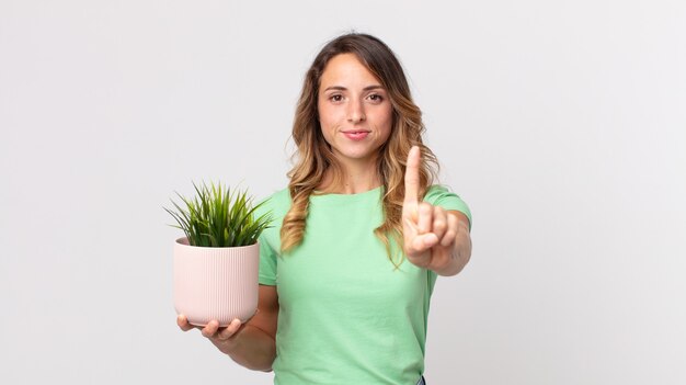 Foto mulher bonita e magra sorrindo com orgulho e confiança fazendo o número um e segurando uma planta decorativa