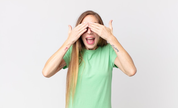 Mulher bonita e magra sorrindo e se sentindo feliz, cobrindo os olhos com as duas mãos e esperando por uma surpresa incrível