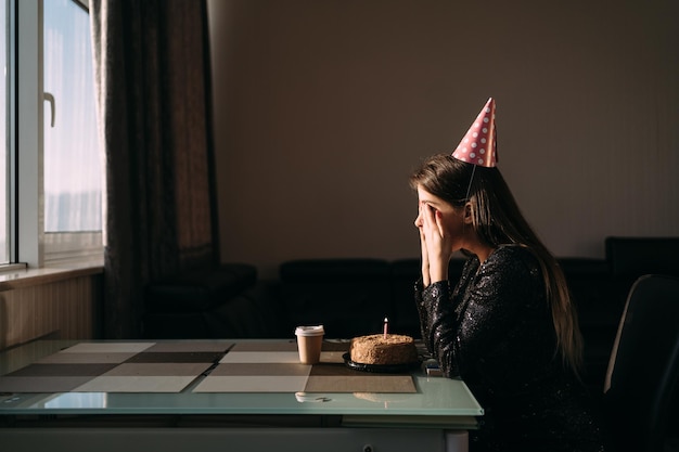 Mulher bonita e pensativa com chapéu de festa olhando para bolo festivo com vela freelancer de mulher solitária comemorando aniversário sem amigos