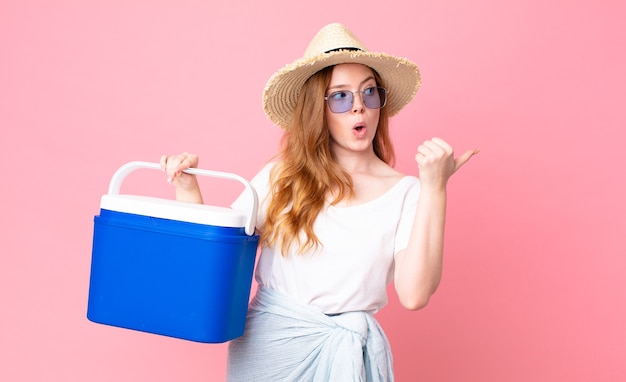 Mulher bonita e ruiva olhando espantada de descrença e segurando uma geladeira portátil de piquenique