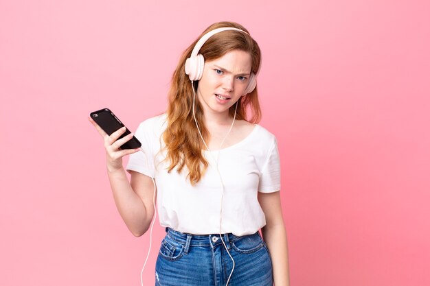 Mulher bonita e ruiva se sentindo confusa e confusa com fones de ouvido e smartphone