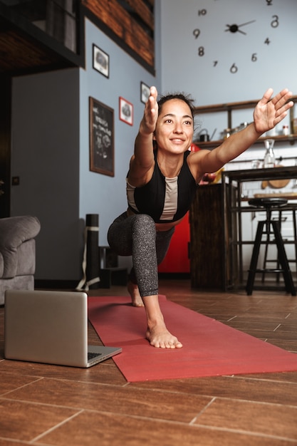 Mulher bonita e saudável fazendo exercícios de ioga enquanto está sentado em uma esteira de ginástica em casa, usando um laptop, fazendo alongamento