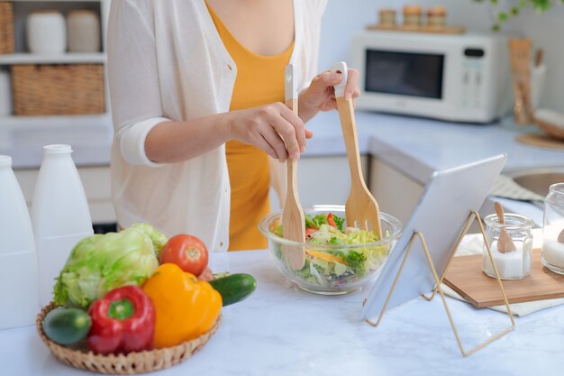 Mulher bonita e sorridente comendo vegetais enquanto prepara uma salada de uma receita no computador tablet em uma cozinha