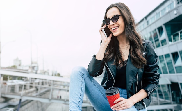 Mulher bonita elegante e moderna com óculos escuros e estilo hippie, use enquanto ela está usando um telefone e se diverte ao ar livre na cidade