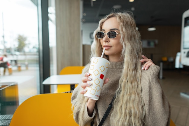 Mulher bonita elegante segurando um copo de papel de cola sentado em um café e bebendo uma bebida