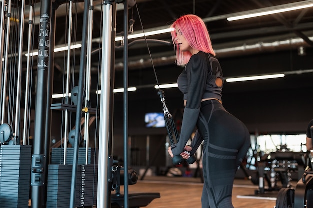 Mulher bonita em fitness com cabelo rosa e um agasalho de treino preto faz exercício e bombeia os músculos no simulador no ginásio