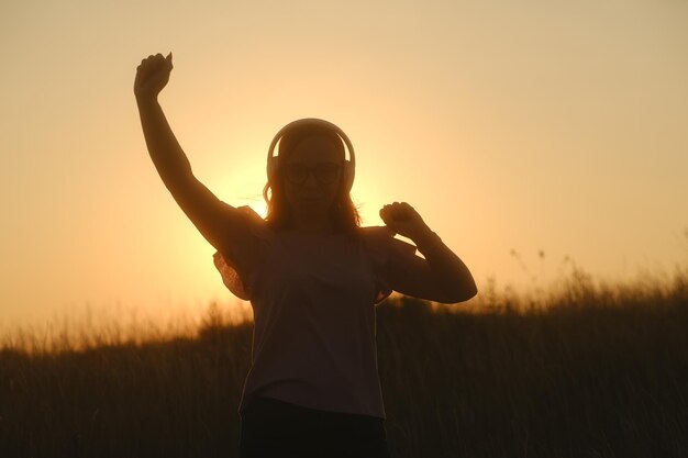 Mulher bonita em fones de ouvido e óculos dançando ao pôr do sol na zona rural ao pôr do sol em roupas casuais