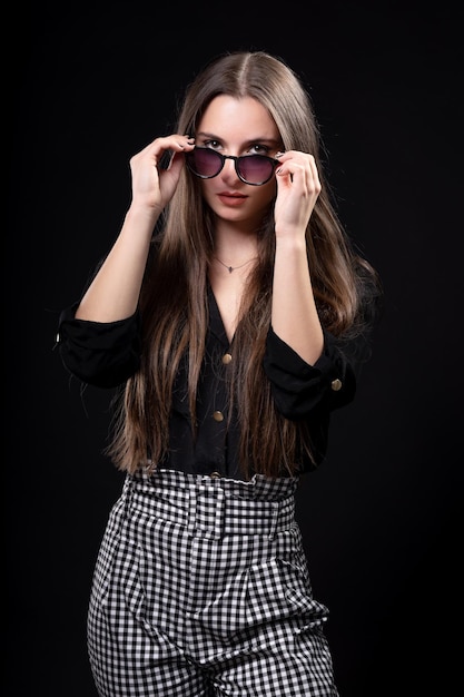 Mulher bonita em fundo preto com camisa preta e calça listrada, colocando seus óculos.