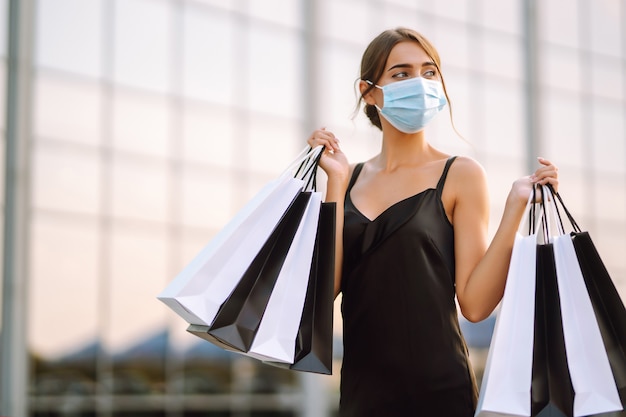 Mulher bonita em máscara médica protetora estéril com sacolas de compras, perto de centro comercial.