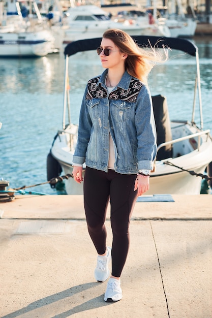 Mulher bonita em óculos de sol redondos e uma jaqueta jeans, à esquerda, longos cabelos loiros em um dia de verão ao ar livre no porto