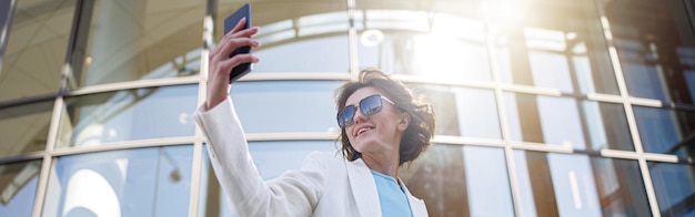 Mulher bonita em roupa da moda fazendo selfie no fundo do edifício moderno