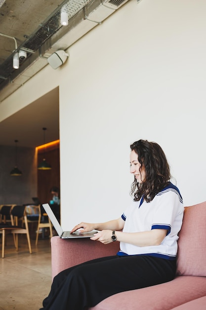 Mulher bonita em roupas casuais sentada em um sofá confortável em um café moderno à mesa com laptop e telefone trabalhando online Trabalho remoto