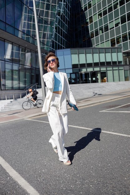 Mulher bonita em roupas da moda atravessa a estrada contra o pano de fundo dos edifícios da cidade