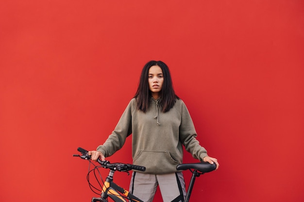 Mulher bonita em roupas esportivas fica em um fundo vermelho com uma bicicleta e posa para a câmera