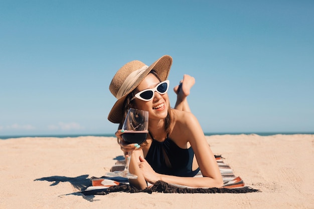 Mulher bonita em trajes de banho sensuais segurando um copo de vinho tinto deitado na praia tropical