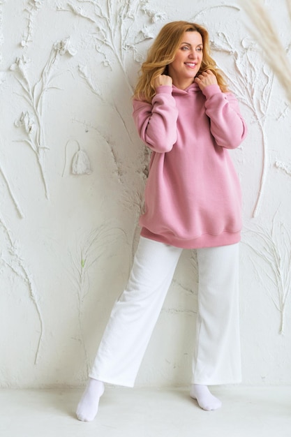 Mulher bonita em um capuz rosa e calça branca posando contra um fundo de uma parede branca. O conceito de roupas bonitas e confortáveis