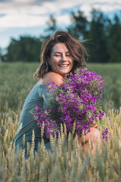 Mulher bonita em um lindo vestido azul e um enorme buquê de flores silvestres roxas ri e se senta no campo