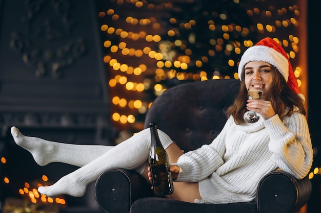 Mulher bonita em um suéter quente, meias e chapéu de Natal, sentada na cadeira com champanhe