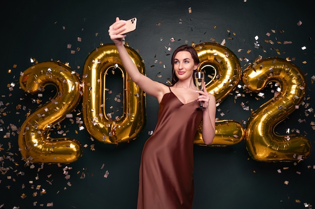 Mulher bonita em um vestido moderno de luxo, fazendo selfie com champanhe na frente de balões dourados isol ...