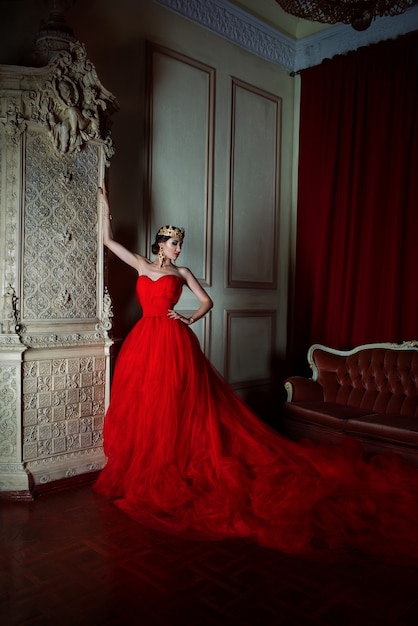 Mulher bonita em um vestido vermelho longo e uma coroa real perto da lareira em um interior luxuoso