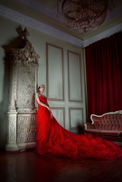 Mulher bonita em um vestido vermelho longo e uma coroa real perto da lareira em um interior luxuoso