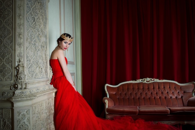 Mulher bonita em um vestido vermelho longo e uma coroa real perto da lareira em um interior luxuoso