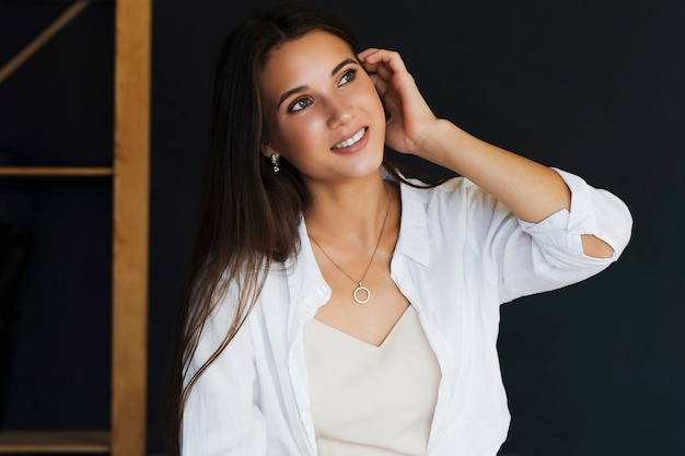 Mulher bonita em uma camisa branca na parede escura. Feche o retrato de uma mulher sorridente com cabelo escuro.