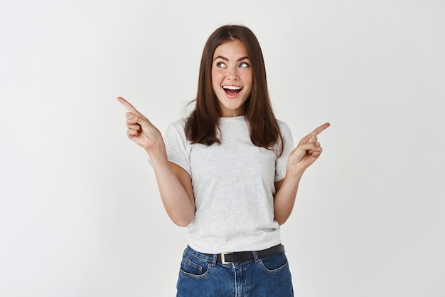 Mulher bonita em uma camiseta casual apontando os dedos para os lados, escolhendo entre duas opções, olhando para a esquerda e sorrindo, em pé sobre uma parede branca.