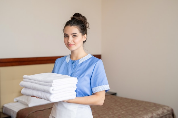 Mulher bonita em uniforme de camareira olhando para você enquanto carrega uma pilha de toalhas brancas macias