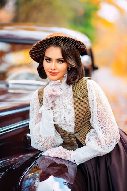 Mulher bonita em vestido vintage, blusa de renda e chapéu com véu em pé perto de carro marrom retrô