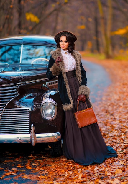 Mulher bonita em vestido vintage, blusa de renda e chapéu com véu em pé perto de carro marrom retrô