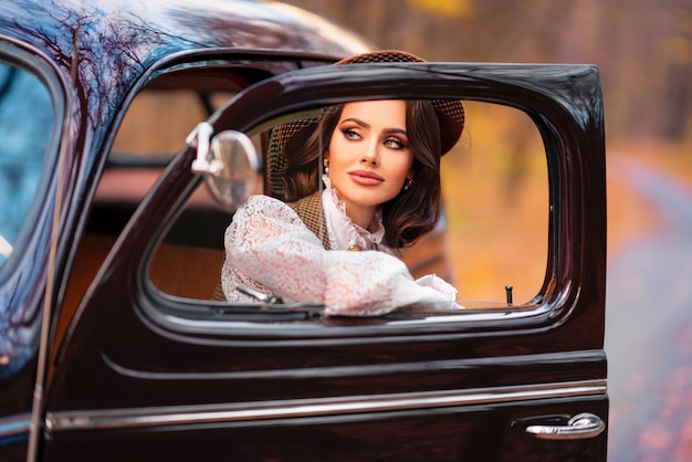 Mulher bonita em vestido vintage, blusa de renda e chapéu com véu em pé perto de carro marrom retrô