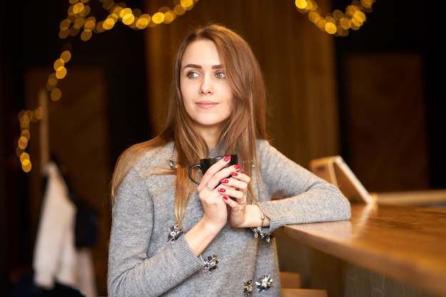Mulher bonita encantadora atraente, com cabelos longos e sorriso amigável localização no café e segurando a xícara de café preta.