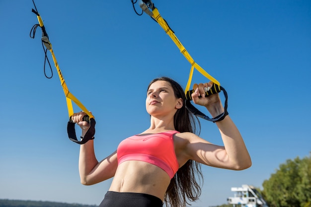 Mulher bonita, exercitando-se com alças de suspensão TRX ao ar livre perto do lago durante o dia. Estilo de vida saudável