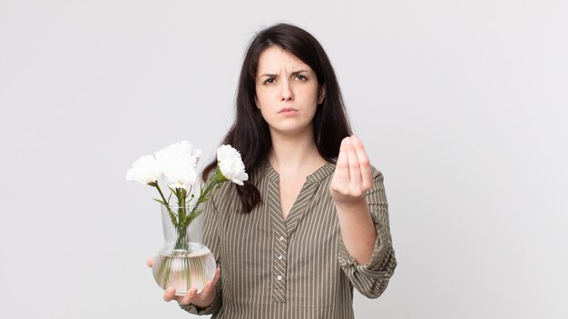 Mulher bonita fazendo capice ou gesto de dinheiro, dizendo para você pagar e segurando flores decorativas. agente assistente com fone de ouvido