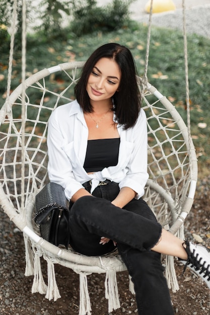 Mulher bonita feliz em roupas de moda casual com jeans preto de camisa branca e bolsa da moda senta e descansa no balanço de malha ao ar livre