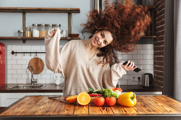 Mulher bonita feliz ouvindo música no celular enquanto cozinha salada de legumes frescos no interior da cozinha em casa