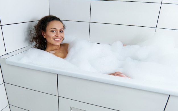 Mulher bonita feliz relaxando em uma banheira de espuma e se divertindo com um banho de espuma