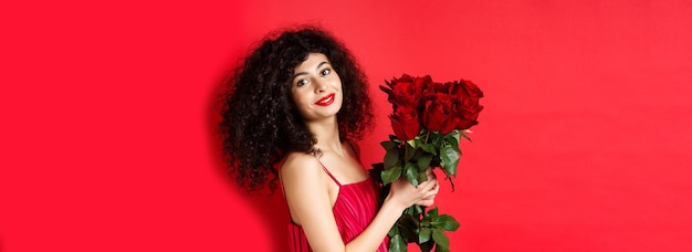 Mulher bonita feliz vestido segurando flores e sorrindo romântico em pé contra o fundo vermelho