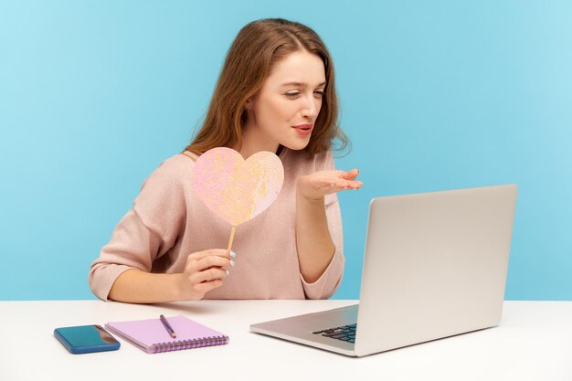 Mulher bonita feminina segurando coração rosa e enviando beijo de ar para a tela do laptop, expressando sentimentos românticos, tendo namoro online em quarentena. tiro de estúdio interno isolado em fundo azul