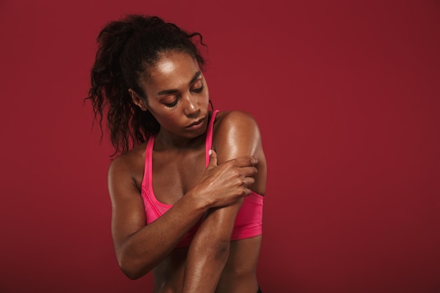 mulher bonita forte jovem africano fitness posando isolado sobre a parede vermelha, tocando seu braço.