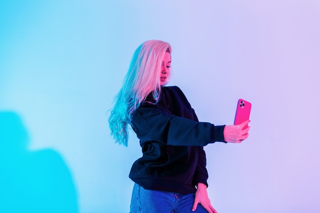 Mulher bonita glamourosa na moda em um moletom preto casual com jeans que faz uma selfie em um smartphone no estúdio em um fundo azul-rosa multicolorido