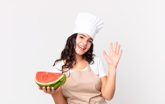 Mulher bonita hispânica chef sorrindo feliz, acenando com a mão, dando as boas-vindas, cumprimentando você e segurando uma melancia