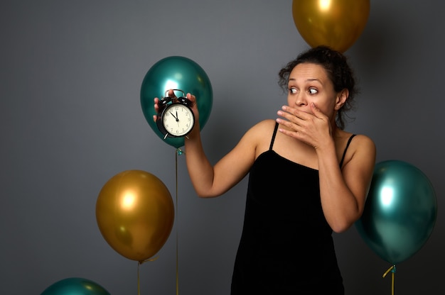 Mulher bonita hispânica com um vestido preto de noite, cobre a boca, surpresa com a hora no despertador, é meia-noite. Isolado sobre um fundo cinza com balões de ar verde dourado Copiar espaço