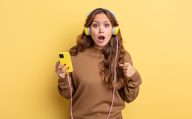 Mulher bonita hispânica parecendo chocada e surpresa com a boca aberta, apontando para si mesma. conceito de fones de ouvido e smartphone