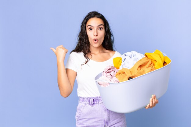 Mulher bonita hispânica parecendo espantada com a descrença e segurando uma cesta de roupas para lavar